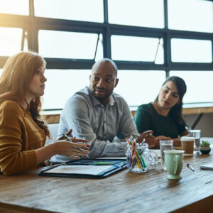 Three professionals sit at a table with pens, cups, and paper in front of them. One is speaking and the other two are looking at her.