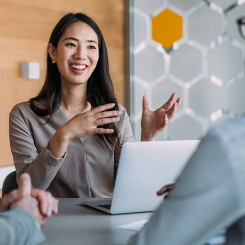 Professional woman in an office environment, sitting at a table with a computer in front of her, speaking to others at the table