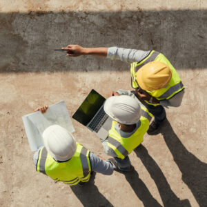 Three people in hi-vis vests and hard hats standing side by side, one with blueprints, one with a laptop, and the other pointing at something in the distance