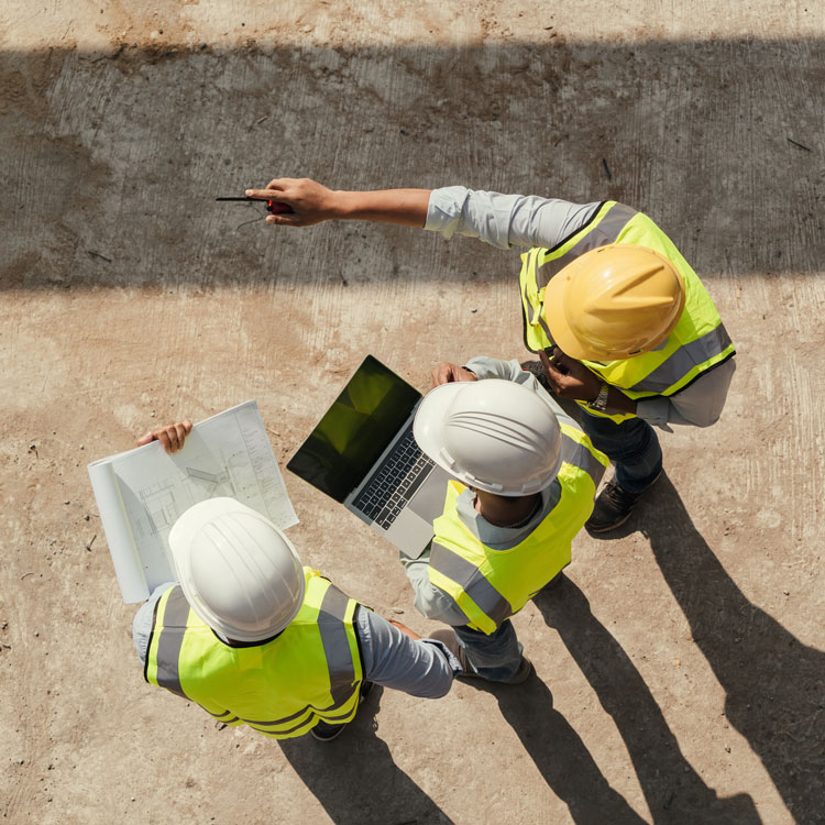 Three people in hi-vis vests and hard hats standing side by side, one with blueprints, one with a laptop, and the other pointing at something in the distance