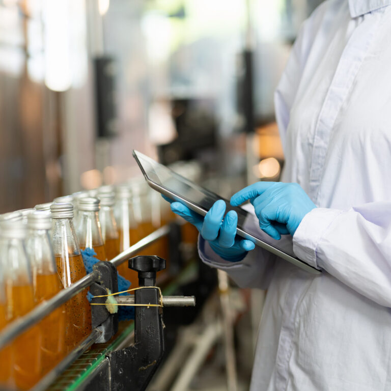 Worker in a lab coat with latex gloves and a digital tablet near a beverage product line